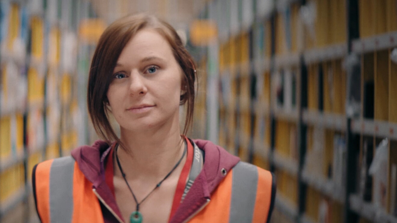 A woman wearing a safety vest stands in a warehouse space.