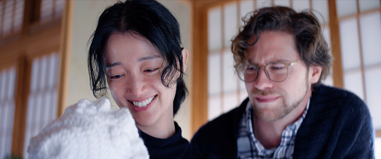 和室の障子を背景に並んだ2人の男女のアップ。左側女性の体の前に、白いかぎ針編み帽子の一部。女性も男性を帽子の中を見つめている。女性は笑顔だが、男性は泣きそうな表情。