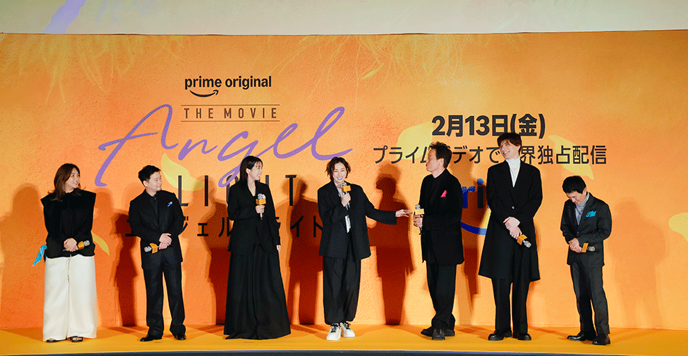 オレンジ色の壁の前、オレンジのステージの上に、黒い服を着て横一列に並んだ7人の人物。右端の人物だけグレーの上下。中央の人物が左手を隣の人物に笑顔で伸ばしている。