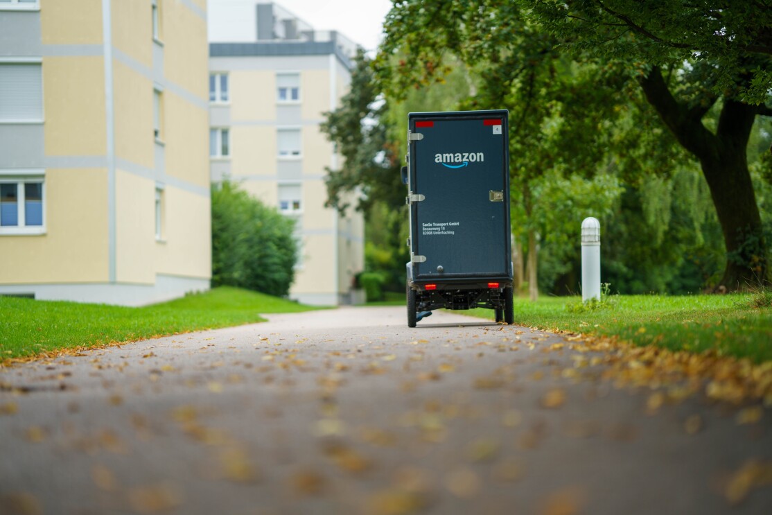 Ein E-Cargo Bike mit Amazon-Logoaufdruck fährt auf einem Fahrradweg. Man sieht links Häuser, recht viel grün, auf dem Weg erstes Herbstlaub.
