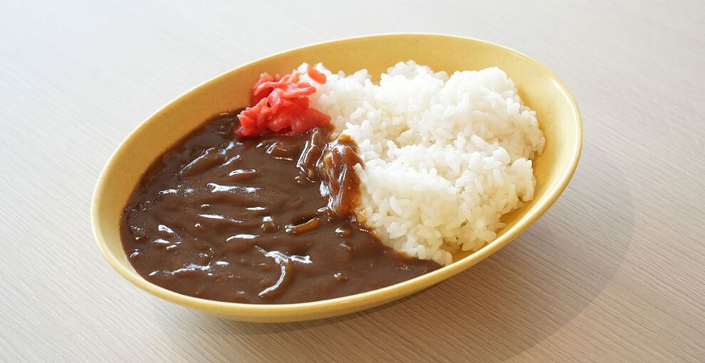 木目の表面に置かれた黄色い楕円形の皿。皿の中にはカレーライスが盛られている。左側がカレールー、右側がライス。ルーとライスの境目に赤い福神漬けが置かれている。