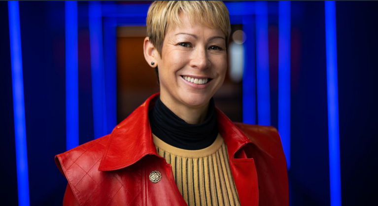 An image of a woman smiling for a headshot in a hallway of blue lights. She is wearing a red jacket and yellow sweater.