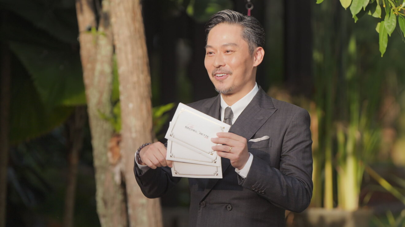 夜の屋外で、南洋の植物を背に立つスーツ姿の男性。体の前で、3つの封筒を持って見せている。
