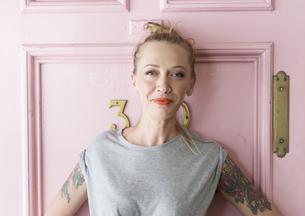 A woman wearing a gray, cropped t-shirt stands in front of a pink door.