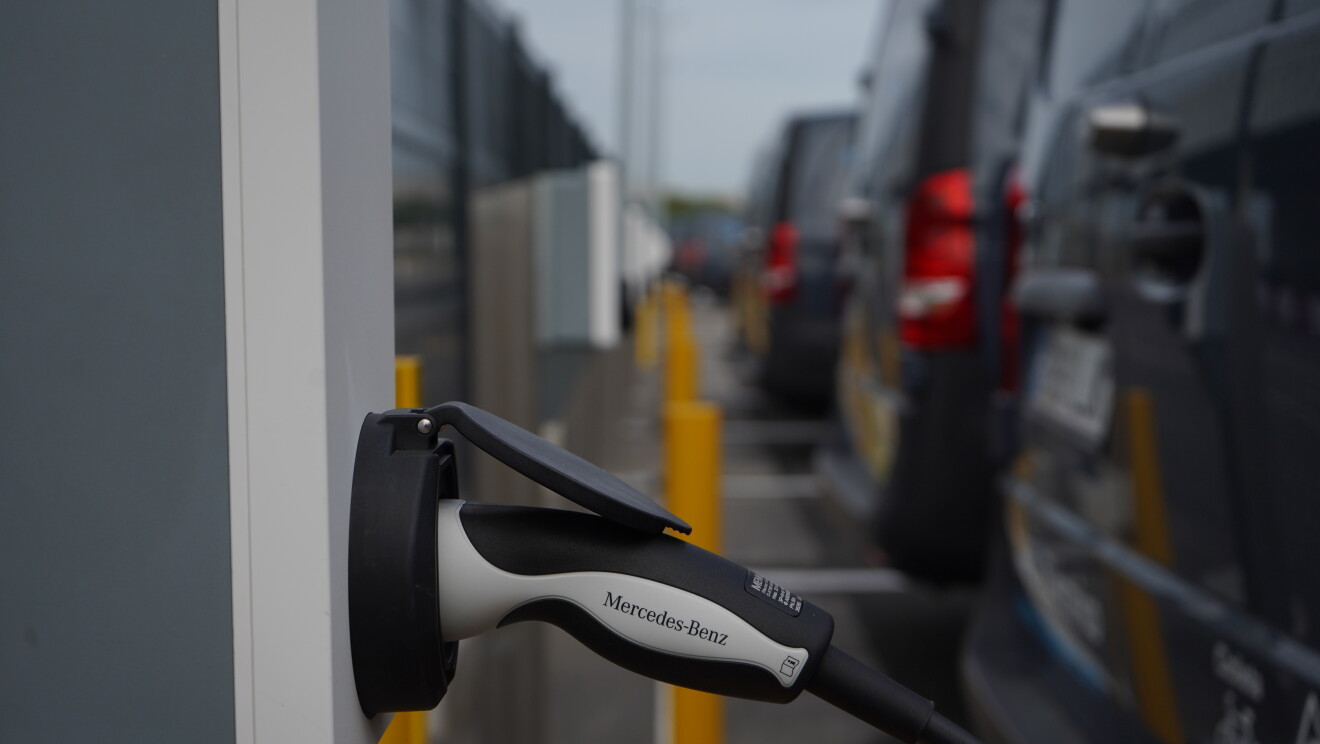 Una Mercedes Benz-Vans en un aparcamiento. Se ve el logo Prime de Amazon y el texto en la puerta 100% Eléctrico Envíos 'Shipment Zero'. En primer plano se ve un cargados de batería que está subministrando electricidad a la furgoneta de al lado que solo se ve un poco su parte trasera. 
