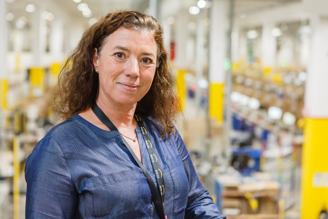 Eine braunhaarige Frau in blauer Bluse in einem Logistikzentrum (Aufnahme vor Pandemie).