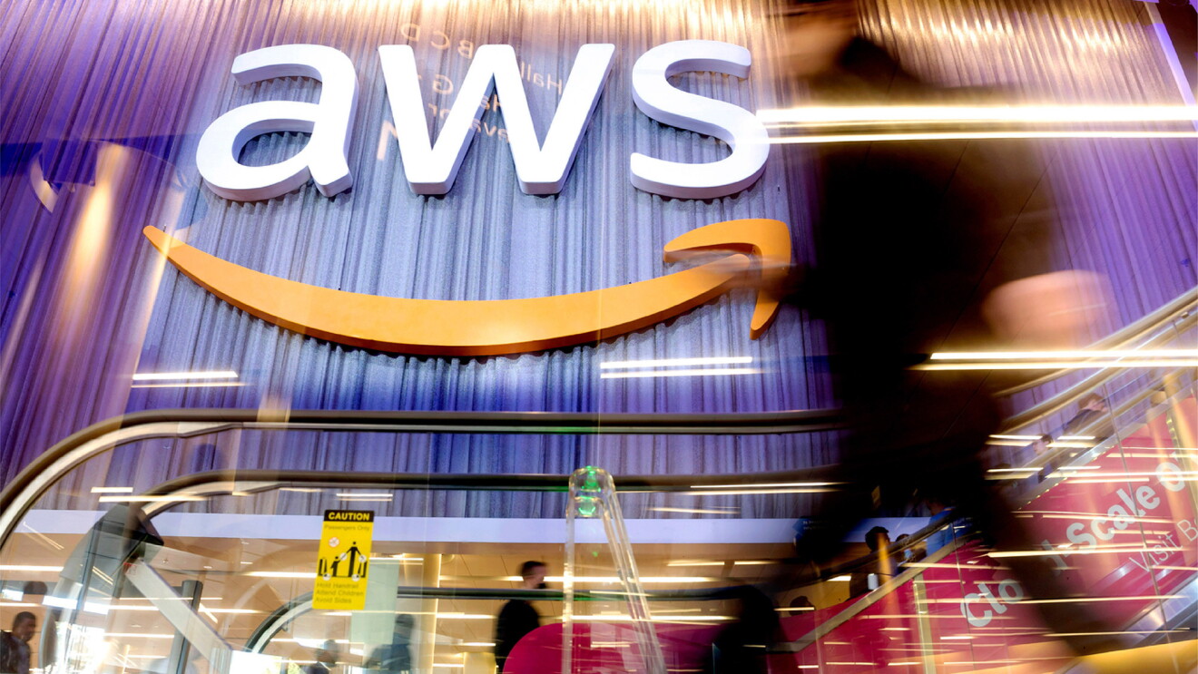 A photo of an AWS logo sign on the side of a conference center wall. There is an escalator handle in the forefront of the photo.