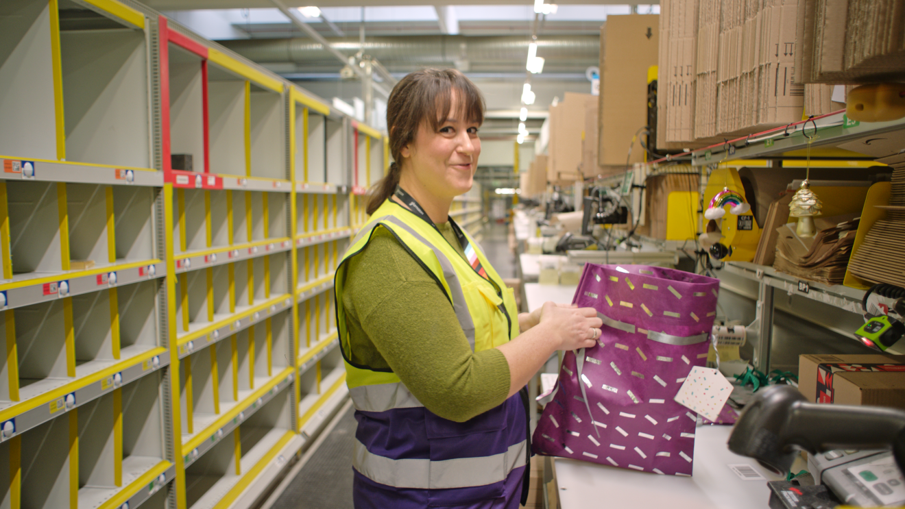 Eine Amazon Mitarbeiterin vor einem Tisch im Amazon Lager: Sie verpackt eine Bestellung als Geschenk.