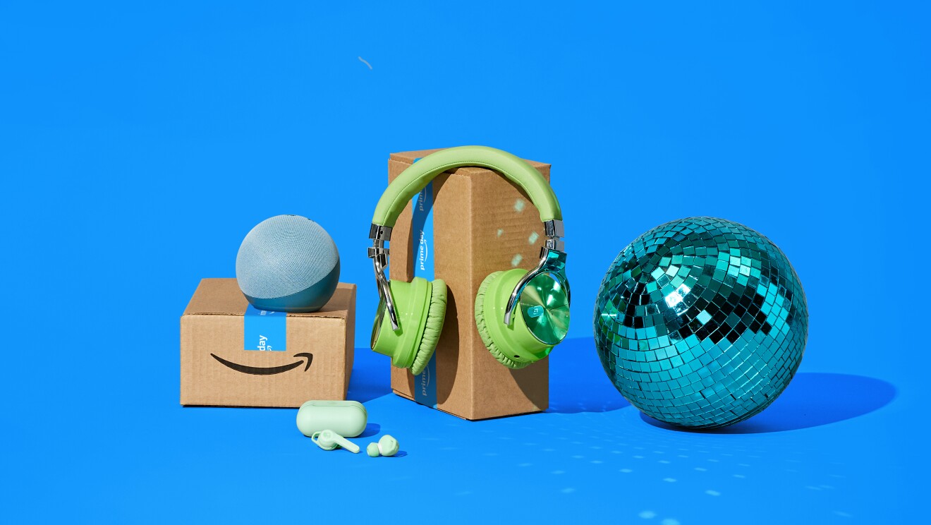 Fotomontaje, sobre un fondo azul, dos cajas de Aamzon, una con dos dispositivos Amazon apoyados, la caja grande con unos auriculares verdes y al lado derecho una bola de fiestas de discoteca.