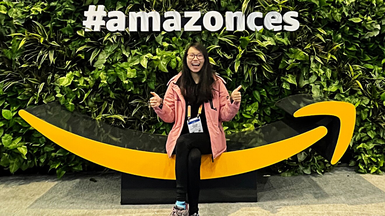 Phoebe Wang smiling to the camera sitting on a big Amazon smile logo with a sign that reads #amazonces behind her. 