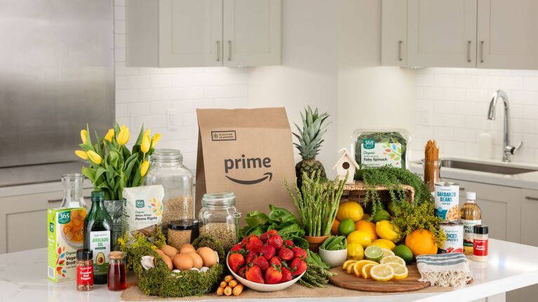 Amazon Prime grocery delivery bag surrounded by fresh produce, flowers, and pantry items on a white kitchen counter