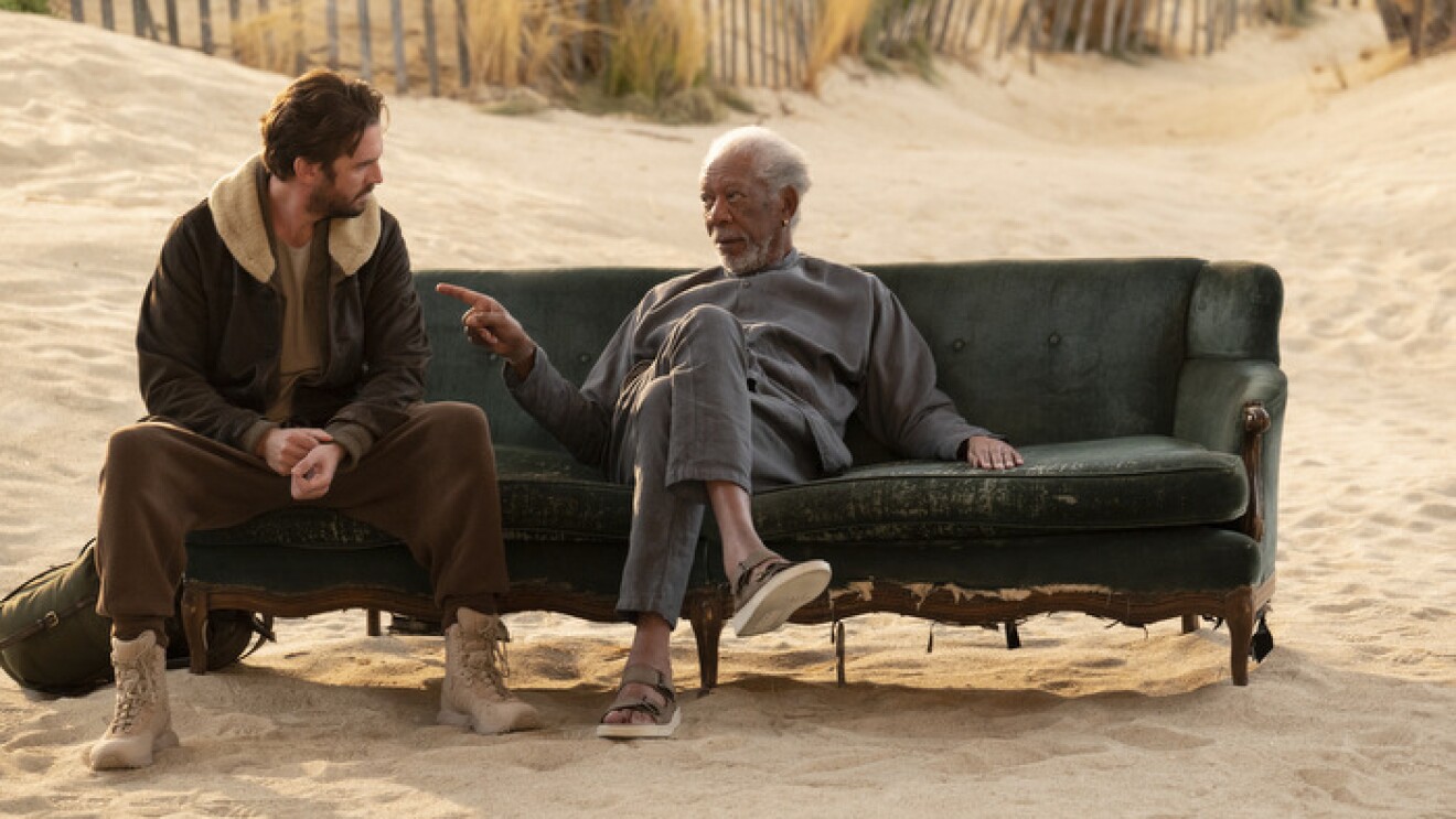 An image of Morgan Freeman and Dan Stevens sitting on a couch on a beach talking to each other. Freeman is pointing at Stevens casually as he speaks.
