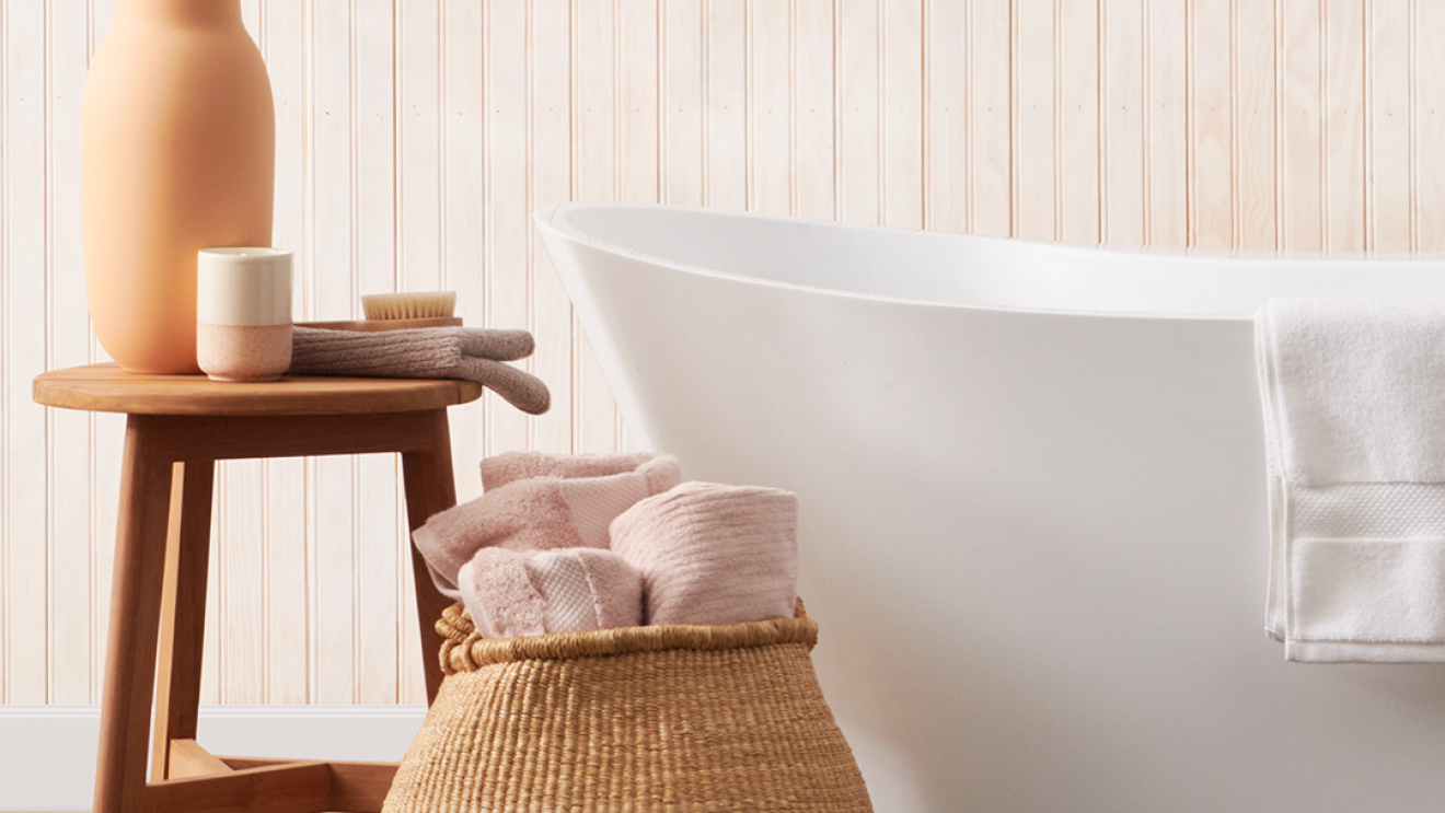 Eine weiße freistehende Badewanne. Daneben steht ein Bastkorb mit Handtüchern und ein kleiner Holztisch mit Schwämmen, Dosen und einer Blumenvase.
