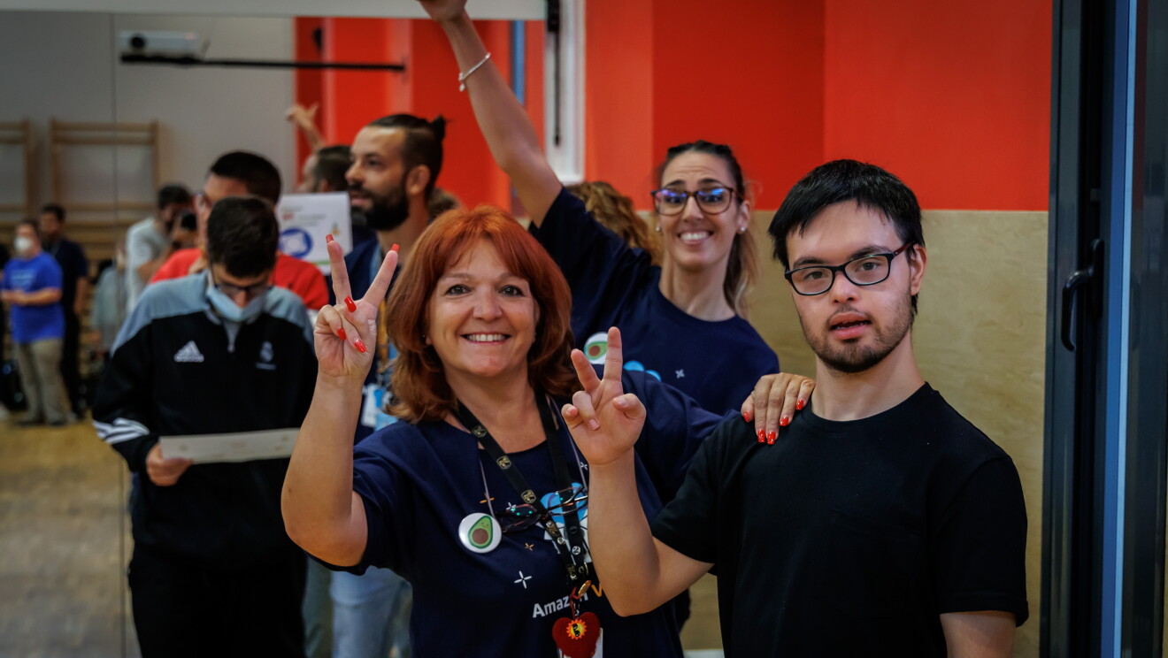 En las instalaciones de Down Madrid en Madrid aparecen 3 personas, 2 trabajadoras de Amazon y 1 participante de la fundación. Los trabajadores de Amazon van con la misma camiseta azul y el badge colgado y los de Down Madrid con deportivas y ropa cómoda. Los tres hacen con la mano izquierda la señal de victoria. 