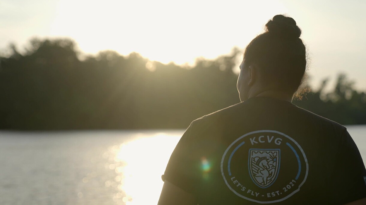 Newton wears a T-shirt that reads KCVG on the back. She peers out over a lake as the sun sets.