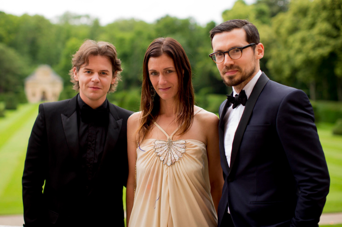 An image of Sally Singer smiling for a photo at a black tie event. There are two men smiling for the photo alongside her.