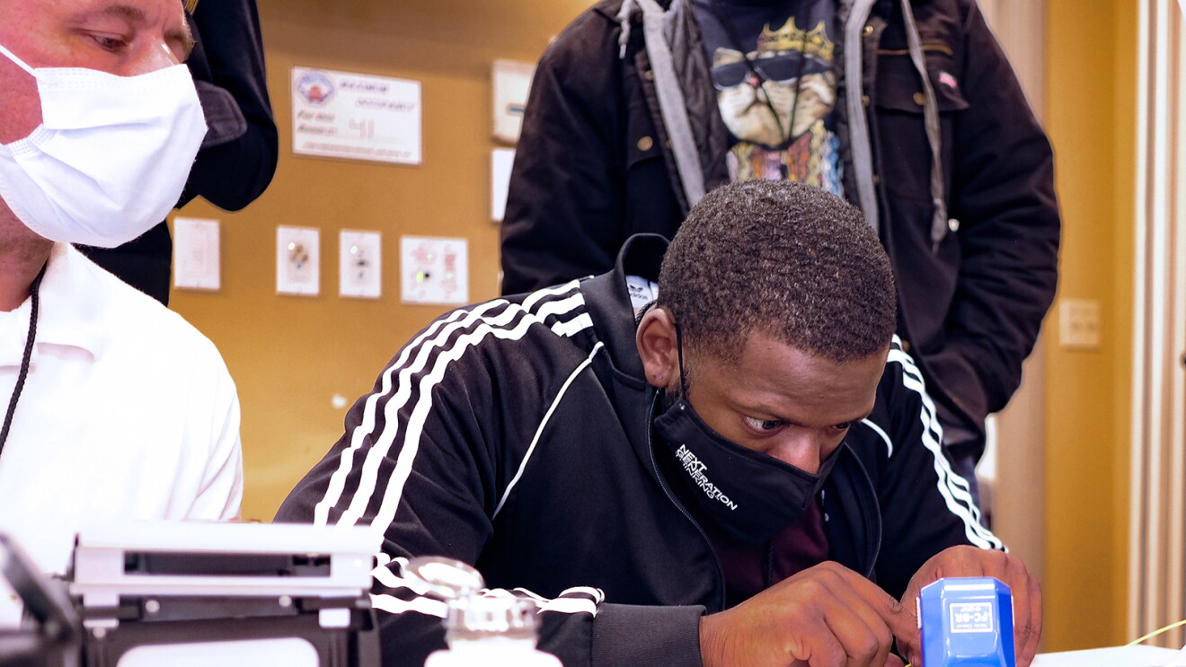 A man is sitting at a table working on a piece of technology hardware. His instructor and fellow students look on as he works. 