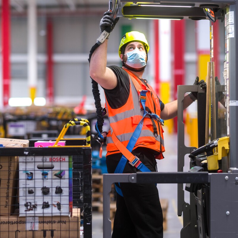 Un empleado de Amazon que se beneficia de la tecnología de arnés de seguridad en altura (en un recogepedidos o un camión industrial motorizado). En un toro hay un empleado de Amazon enganchando su arnés a la parte superior de este. Con la otra mano se sujeta a la barandilla del toro que está alzada. El toro es de color gris y amarillo y el empleado va con unos pantalones negros y una camiseta negra también de manga corta, un chaleco naranja, el arnés, una mascarilla quirúrgica y un casco de color amarillo. El toro tiene un palet agarrado con cajas blancas y maroones. De fondo se ve más material en contenedores.