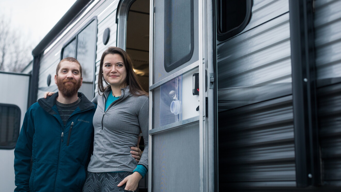 A man and woman stand just outside the door of a silver and gray RV.