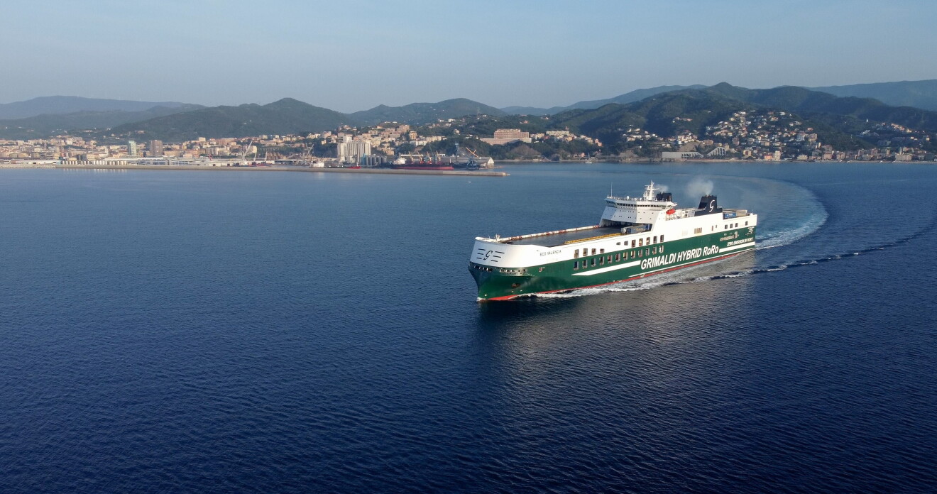 Saliendo de un puerto de una ciudad italiana, se ve, en primer plano un barco blanco y azul de transporte marítimo de camiones llenos de mercancía con las letras Grimaldi Hybro. El mar es de color azul oscuro, el cielo es azul y el pueblo del fondo está entre montañas. 