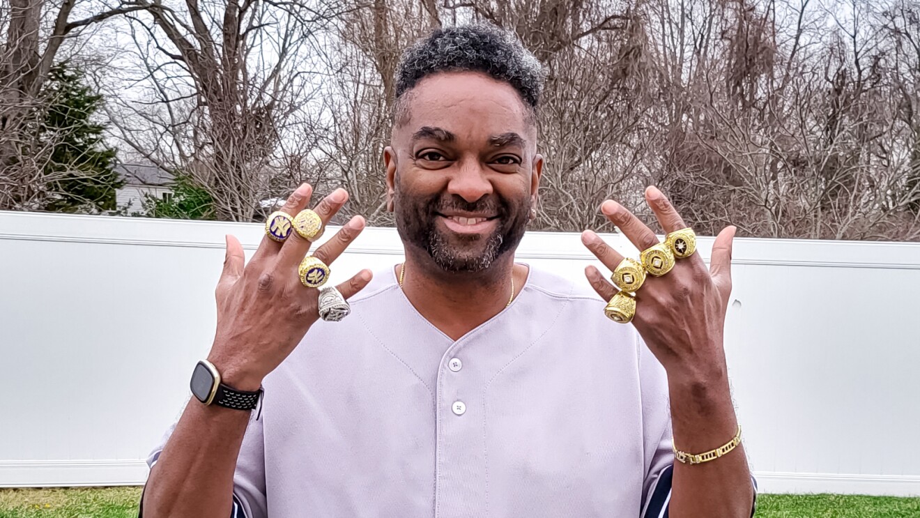 An image of a man smiling for a photo in his backyard. He is holding up both hands to show the championship rings on each of his fingers.