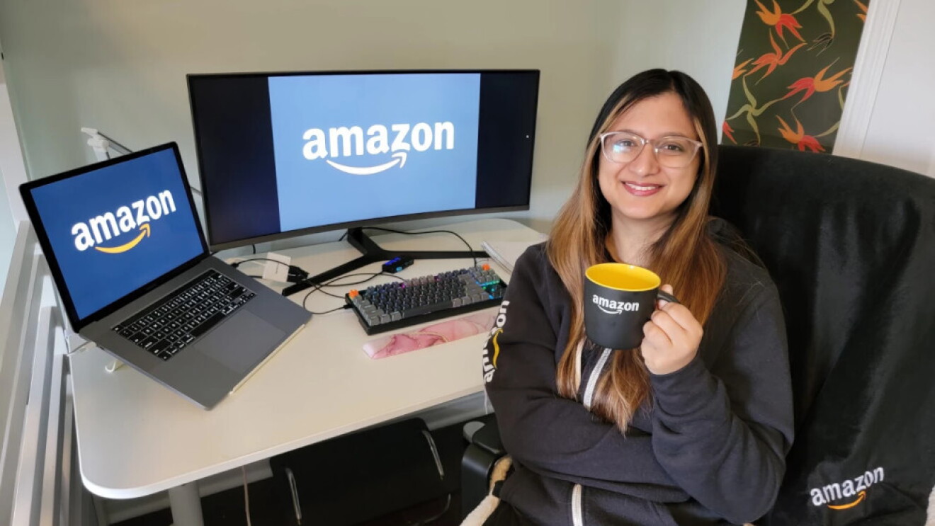 InternDay. Una mujer de unos 25 años con el pelo largo de color negro y mechas de color marrón. Lleva una sudadera de Amazon y está agarrando con la mano izquierda una taza de color negro por fuera y amarilla por dentro con el logo de Amazon en color blanco. Está sentada en una silla delante de su mesa y dándole la espalda. En la mesa hay un ordenador portátil conectado a un display port y tiene una pantalla muy grande y un teclado. En las dos pantallas aparece el logo de Amazon. En la parte izquierda hay una barandilla de hierro blanca y en la derecha un cuadro rectangular vertical con un estampado de hojas de color rojo, naranja y verde. 