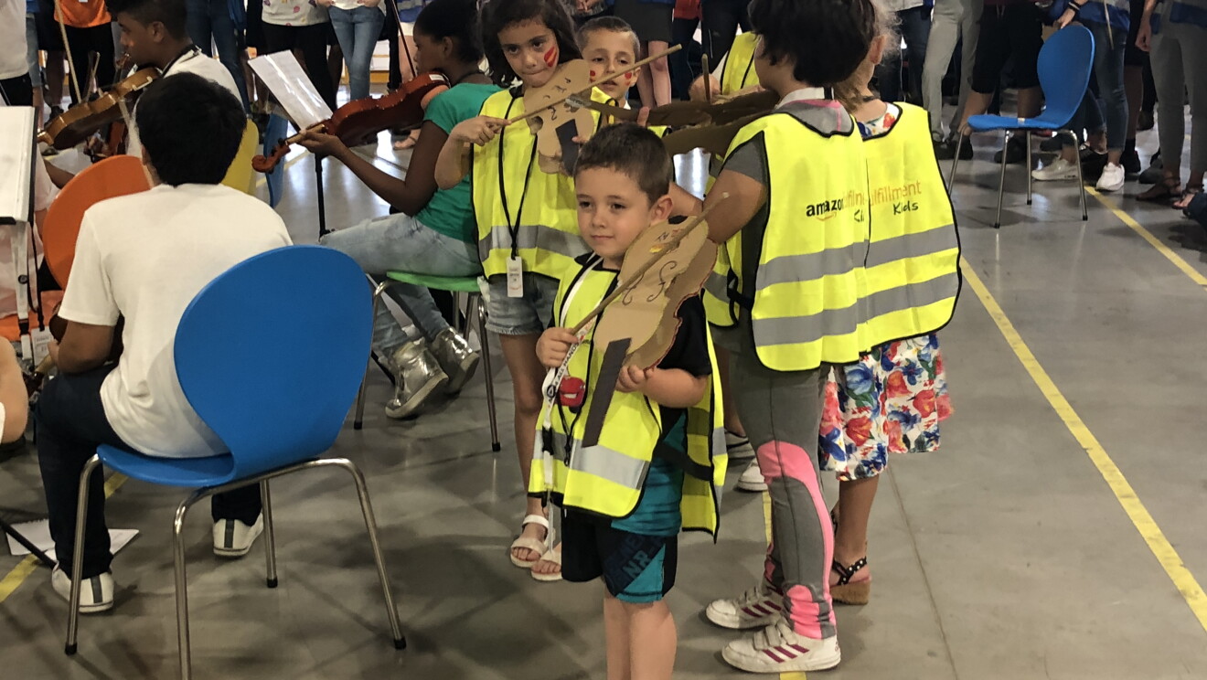 La camerata de Acción Social por la Música se prepara para empezar el concierto en el centro logístico de San Fernando de Henares. En el fondo y de pie, los asociados. En sillas de colores y de espaldas, los componentes de la camerata. Y en primer plano, seis niños, con chalecos e instrumentos de cartón.