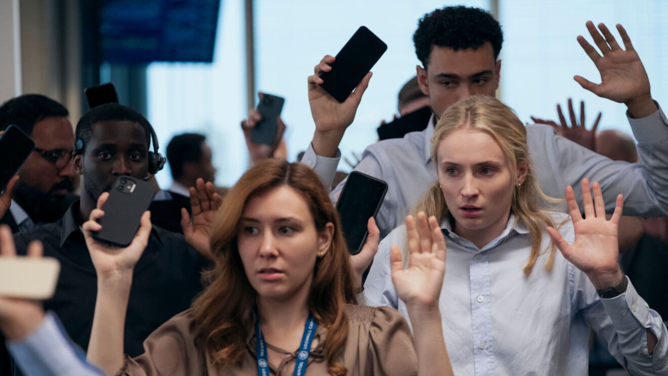 Scene from Steal with office workers holding hands up with smartphones in-hand and fearful expressions