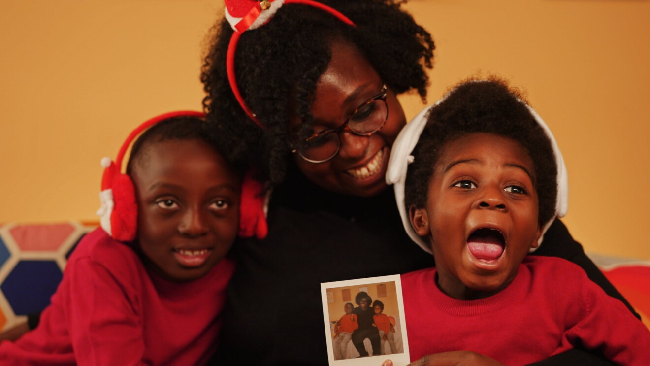 Una mamma con gli occhiali abbraccia i suoi due bambini su un divano. I tre sono vestiti di rosso e hanno cappelli natalizi e paraorecchie a forma di babbo natale. 