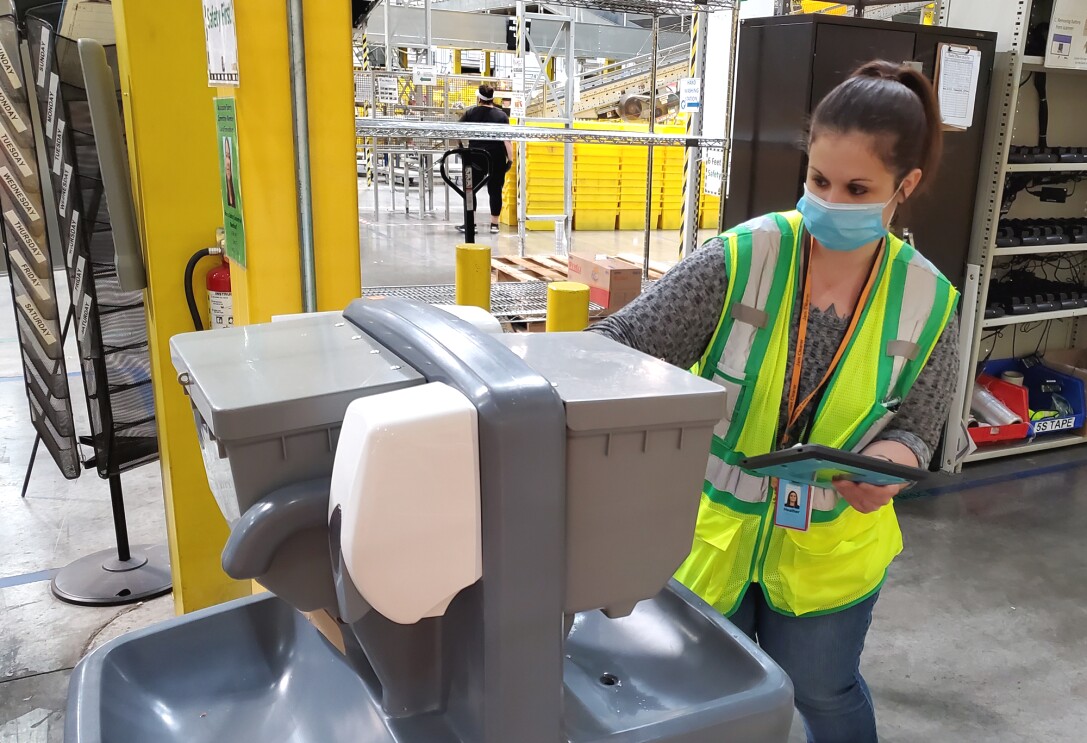 A woman wearing a safety vest and mask inspects a hand-washing station.