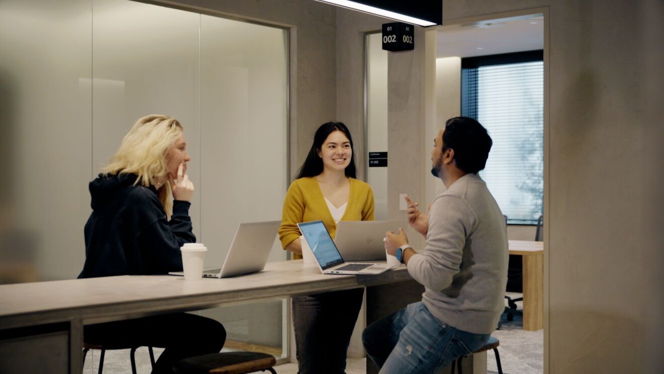 黒髪の女性と金髪の女性とグレーのセーターを着た男性が会議室で談笑している