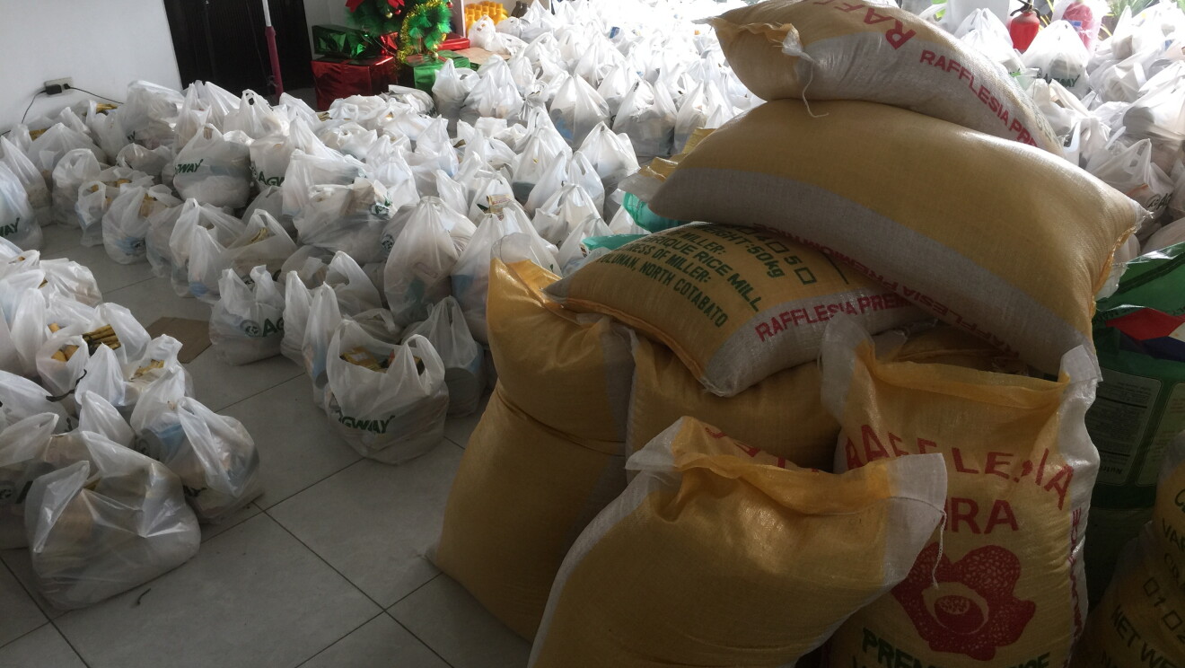 A room full of plastic bags filled food. Gunny sacks filled with rice are stacked to the side of the room.