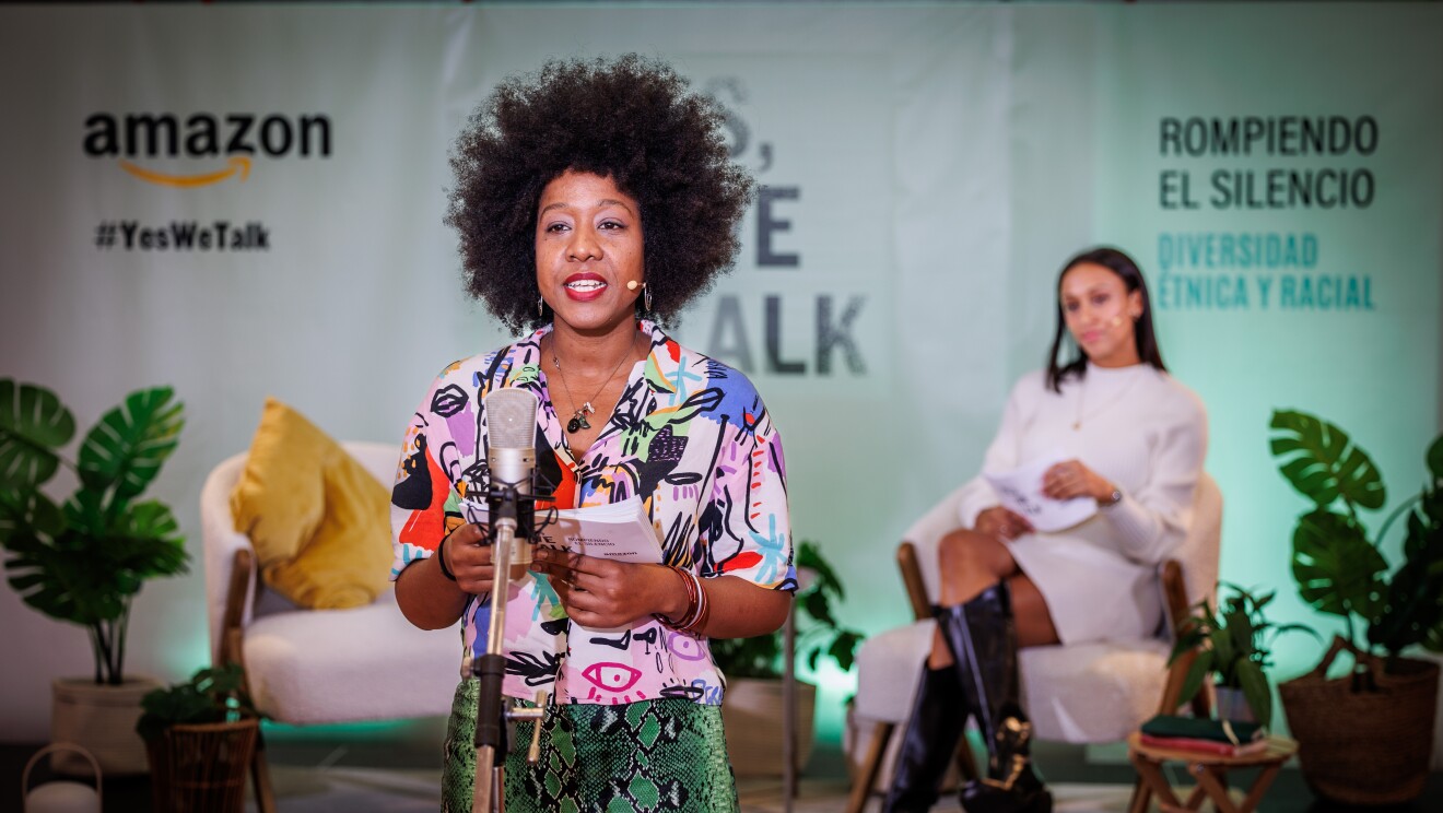Dayan en primer plano y hablando delante de un micrófono. Dayan va vestida con una camisa estampada con dibujos que parecen garabatos de colores de niños y una falta corta estampada con imitación de piel de serpiente de color verde. La deportista Ana Peleteiro, en segundo plano y desenfocada, está  sentada en unas butaca blanca, con las piernas cruzdas. Ana va con un vestido de punto de manga larga, también corto. Las dos son mujeres afrodescendientes. De fondo está el lema de la campaña Yes, we talk. Las protagonistas mirs fíjamente a cámara y la pared de color verde.