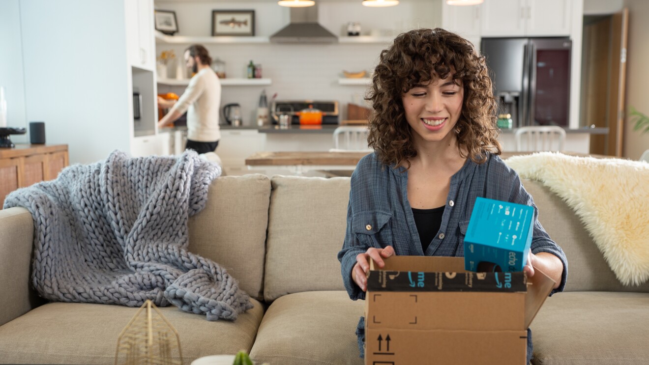 An Amazon customer smiles as they unbox a package on their couch.