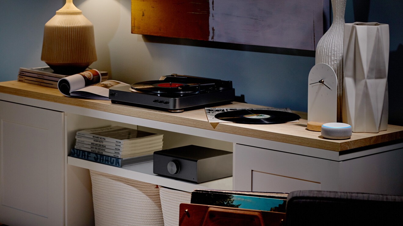 一个回音链接放大器在客厅设置。周围是杂志、唱片、唱机、台灯、装饰品和Echo Dot设备。
