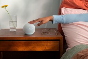 An image of a wooden night stand and with an arm touching an Echo device from a bed. There's a pair of glasses and a vase with a flower next to the device. 