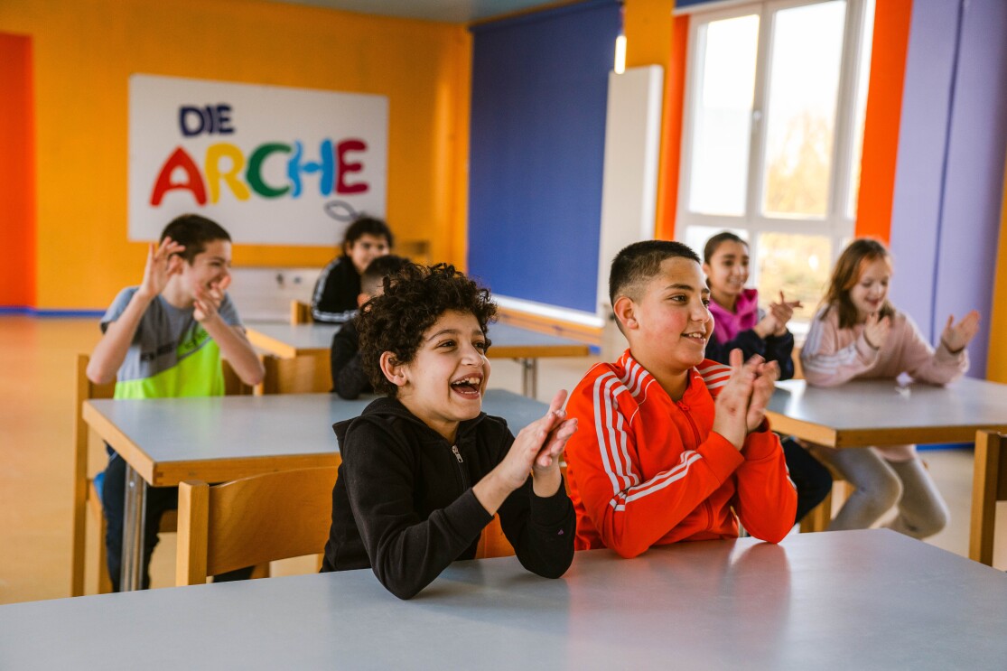 Kinder sitzen in einem Klassenzimmer, jeweils zu zweit an einem Tisch, sie schauen nach vorne und haben Spaß, lachen, klatschen in die Hände. Im Hintergrund hängt das Logo der Organisation "Die Arche".