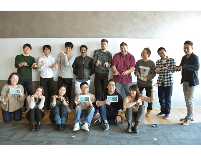 白とグレーの壁を背に集合写真を撮る15人。前列6人、後列9人。キーボード操作のポーズをする人が12人、手に持ったカードを見せる人が3人。
