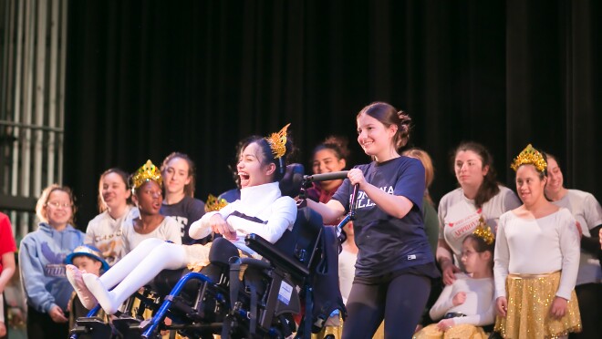 一名舞蹈演员戴着头饰坐在轮椅上。一个十几岁的女孩背后的轮椅,助攻的表现。在后台其他舞者和助手。