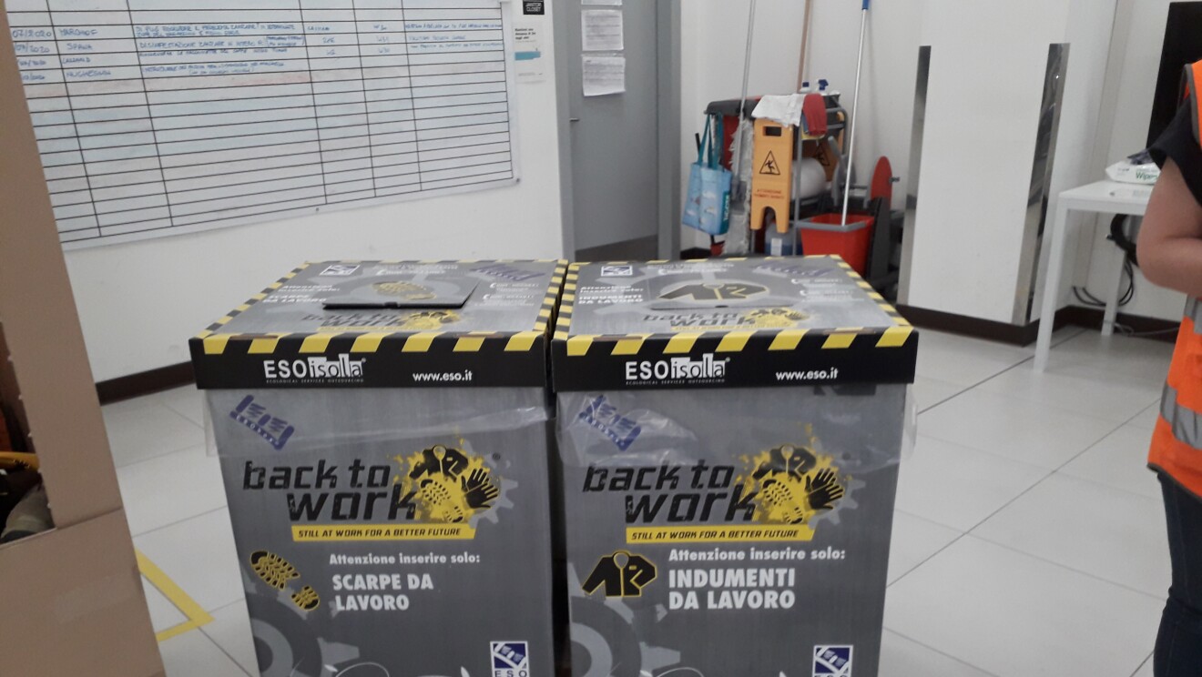 Due scatole di cartone poggiate su un pallet fungono da cestino per inserire indumenti e scarpe da lavoro che verranno poi riciclati. Sulle scatole è scritto 'Back to work'