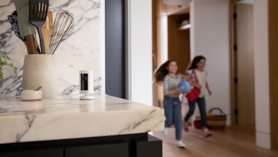 En la parte izquierda de la foto el mármol de la cocina de color blanco y negro con una cámara de seguridad compacta, alimentación por cable, HD, comunicación bidireccional, compatible con Alexa. En el mármol también hay un salero y un recipiente con utensilios de cocina. En la parte izquierda de la foto dos niñas corriendo. Las dos tienen el pelo largo y van vestidas con jeans y deportivas. Una lleva un balón de color azul y la otra un mochila de color rojo. Se ve un mueble de entrar y una habitación con la puerta abierta.