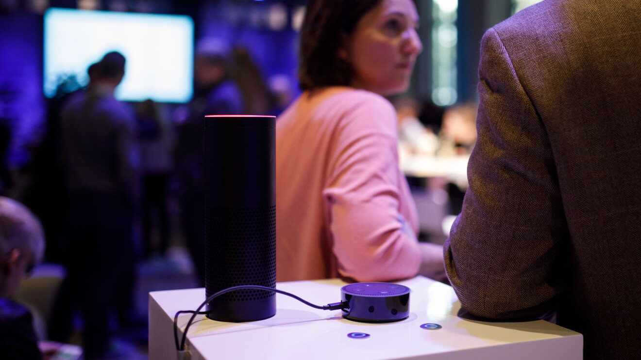 Ein Amazon Echo steht auf dem Tisch. Darum stehen Menschen, die sich unterhalten.