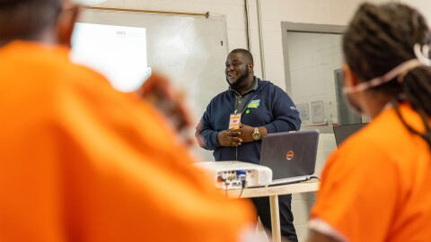 A photo of a cloud computing program instructor presenting in front of a laptop device and projector, with two inmates in the foreground.