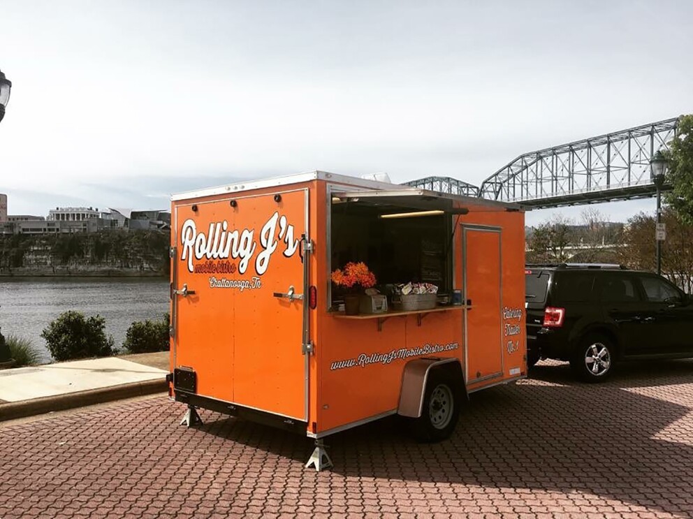 An orange food truck parked in front of the Tennessee River. The food truck has "Rolling J's" branding on it, and is parked on a street made of pavers.