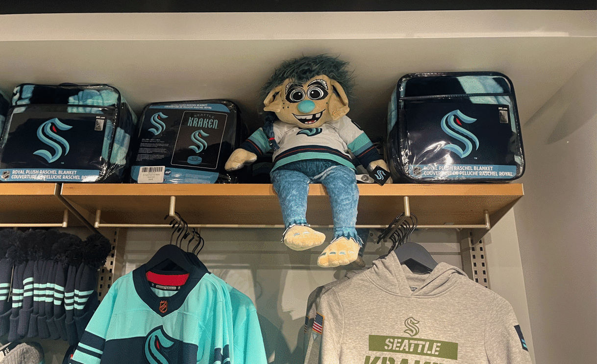 An image of a shelf of merchandise in the Seattle Kraken store. There is a plush toy of the troll mascot, Buoy sitting on the shelf in a Seattle Kraken jersey.