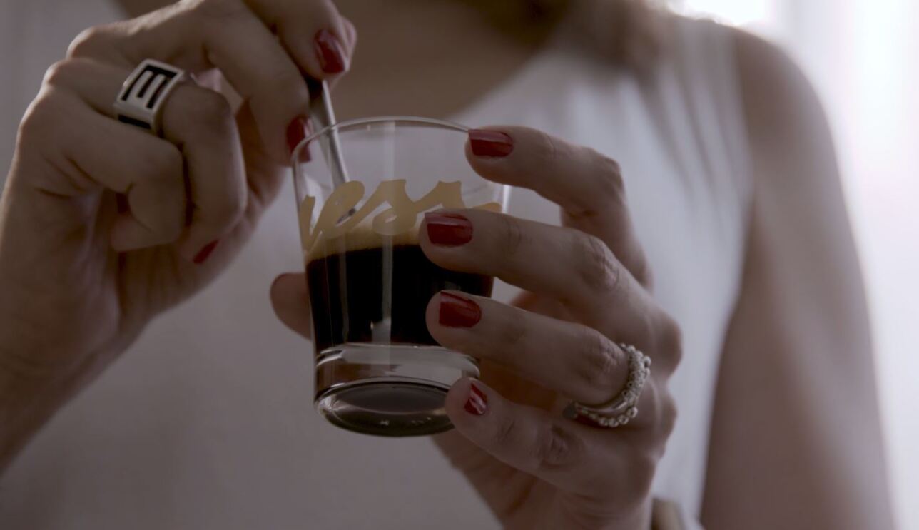 Primo piano di due mani femminili che reggono una tazzina di caffè in vetro. Sulla tazzina si intravede il logo 'Yespresso'. La donna, di cui non si vede il volto, indossa una canotta bianca, ha due anelli su entrambi gli anulari e lo smalto rosso. La sua mano sinistra regge la tazzina, mentre con la destra gira il caffè con un cucchiaino.