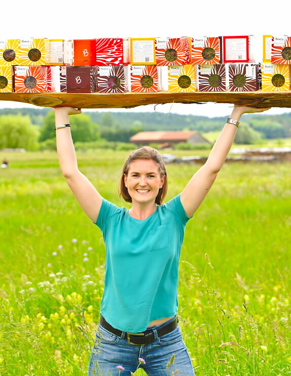 Eine Frau mit braunen Haaren und einem grünen T-shirt steht in einer grünen Wiese und hält ein großes Tablett mit Nudelpackungen über ihrem Kopf.
