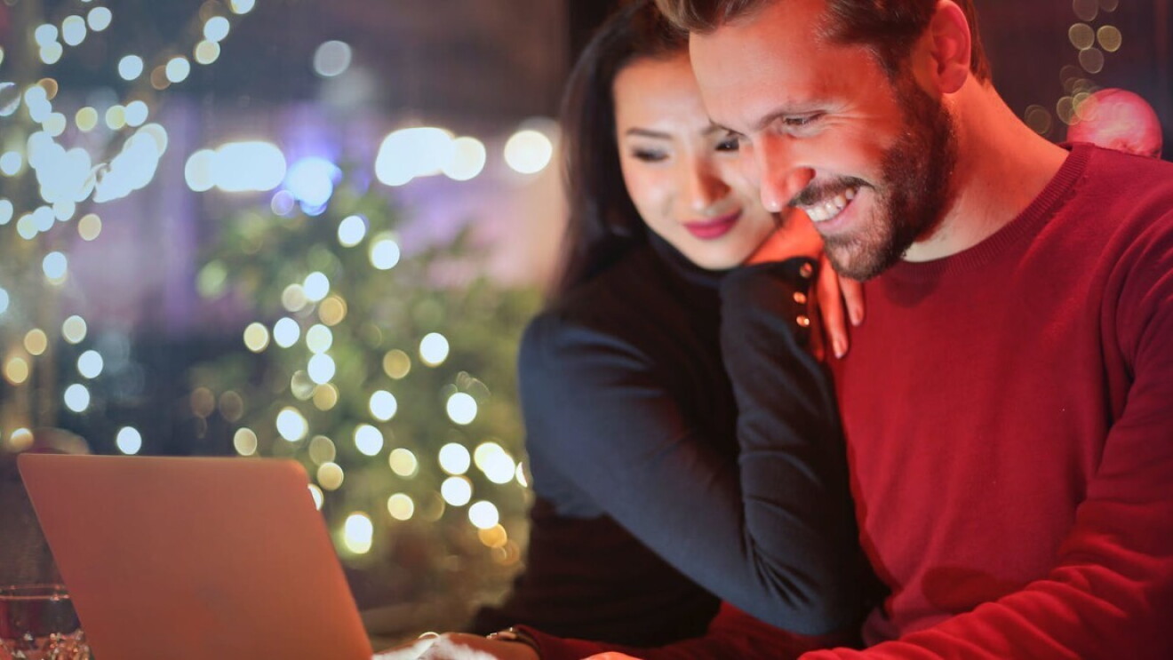 Ein Mann und eine Frau schauen lächelnd in einen hellgrauen Laptop. Im Hintergrund sind verschwommen Weihnachtsbäume und Lichter zu sehen.