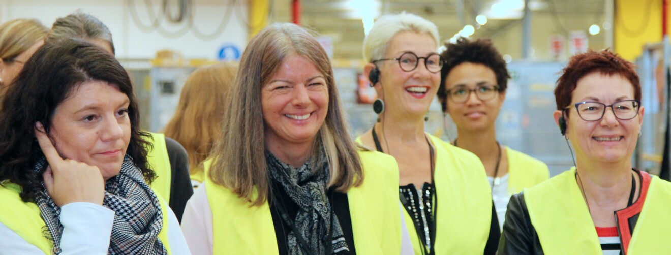 Mehrere Frauen mittleren Alters in gelben Sicherheitswesten stehen hintereinander. Im Hintergrund sind man die Regale des Logistikzentrums.