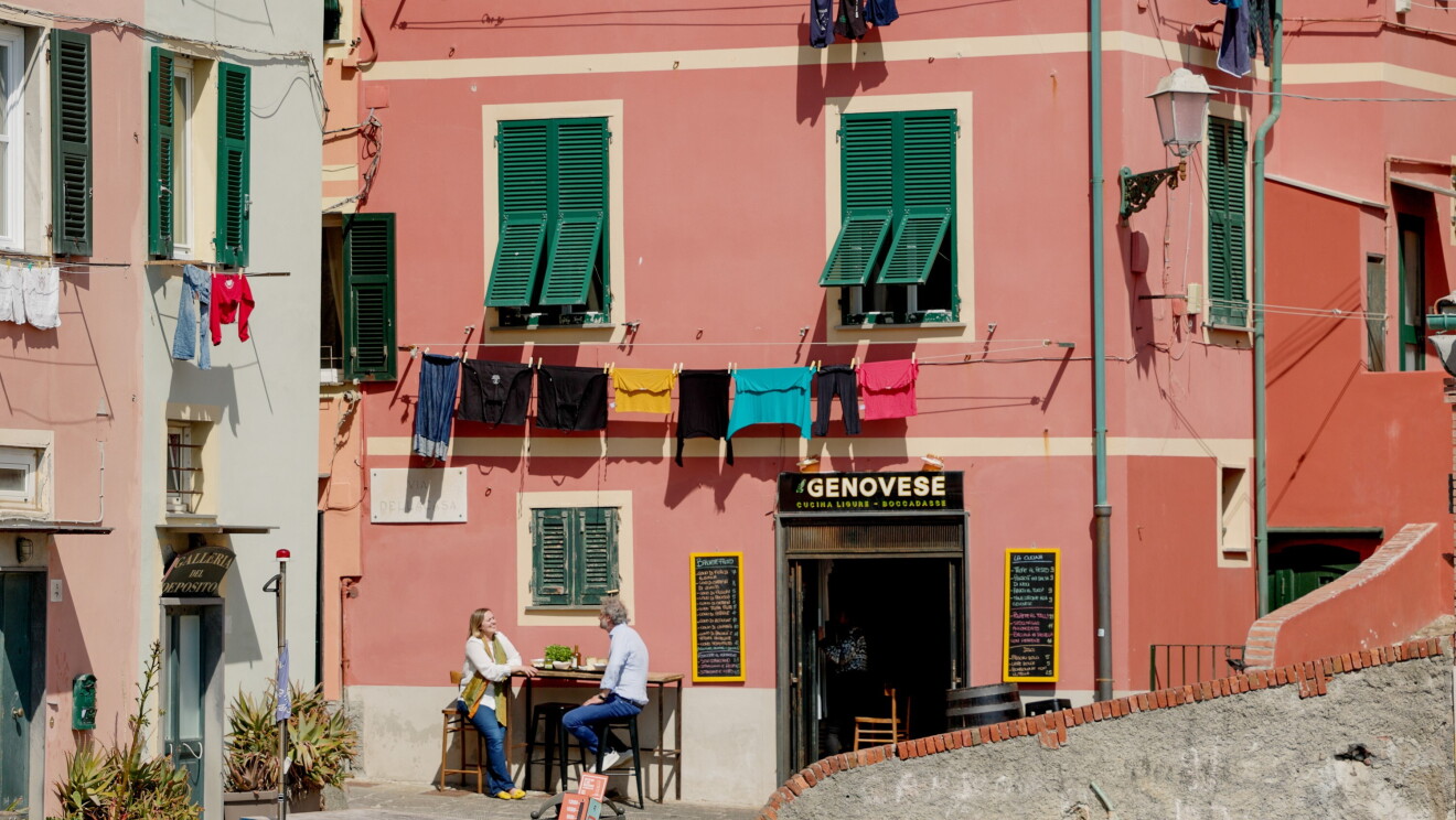 Se ve el centro de un pueblo de Italia, con las fachas de color rosa, un cartel negro con letras blancas que pone Genovese, es un restaurante.  Sabemos que es Génova y que en una mesa alta y dos sillas que están en la parte izquierda de la puerta del restaurante los propietarios de la fábrica de pesto, Pesto Rossi. En las dos fachas de casa que se ven hay ropa tendida. 